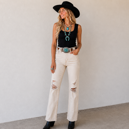 Woman wearing a black top, white jeans, and a black hat with a plain background